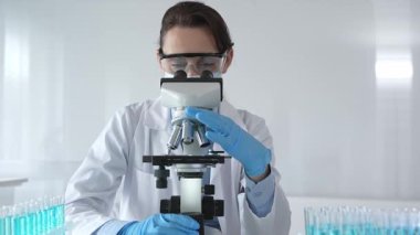 Koruyucu gözlük takan ve maske takan kadın bilim adamı mikroskop kullanarak laboratuvarda numuneleri analiz ediyor mavi sıvı test tüpleriyle çevrili yeni ilaç veya aşıları araştırıyor.
