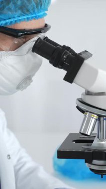 Mavi koruyucu giysiler giyen kadın bilim adamı mikroskop kullanarak laboratuvar ortamında örnekler inceleyerek bilimsel araştırma ve analiz yapıyor. Tıp ve bilim konsepti.