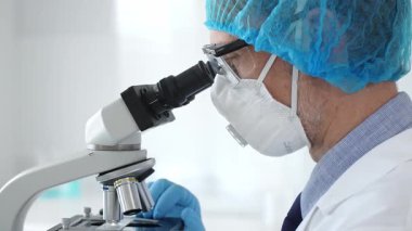 Koruyucu ekipman takan erkek bilim adamı mikroskop kaydırağını inceliyor, steril laboratuvar ortamında araştırma örneklerini analiz ediyor. Sağlık ve bilim konsepti.