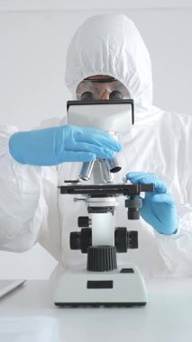 Tehlikeli madde giysisi giyen tıp virolojisti laboratuvarda mikroskopla çalışıyor. Yeni virüsler için potansiyel tedavileri araştırırken mavi sıvı örneklerini inceliyor.