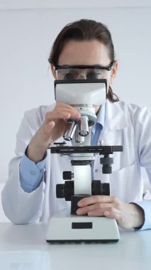 Beyaz laboratuvar önlüğü giyen bilim araştırmacısı mikroskop ile numune inceliyor, test tüpleriyle steril laboratuvar ortamında biyolojik araştırmalar yapıyor.