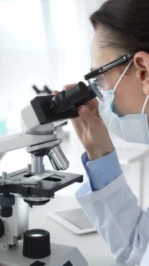 Koruyucu maske takan ve gözlük takan kadın bilim adamı mikroskopla numuneleri dikkatle inceliyor ve modern laboratuvarda araştırma yapıyor. Tıp ve bilim konsepti.