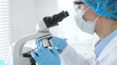 Araştırma örneğini mikroskop üzerinden inceleyen kadın bilim adamı tıbbi araştırma için koruyucu laboratuvar malzemesi giyiyor. Laboratuvar ve bilim konsepti.
