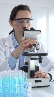 Beyaz laboratuvar önlüğü giyen bilim araştırmacısı mikroskop ile numune inceliyor, test tüpleriyle steril laboratuvar ortamında biyolojik araştırmalar yapıyor.