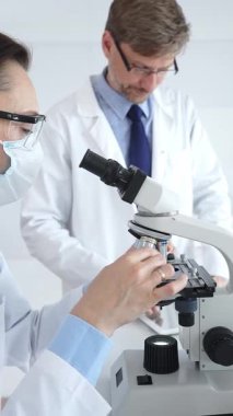 Koruyucu gözlük ve maske takan kadın bilim adamı, laboratuvarda tıbbi araştırma sırasında mikroskoptan bakıyor ve erkek meslektaşı arka planda çalışıyor. Tıp ve Bilim