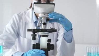 Mikroskobu ayarlayan, laboratuardaki örnekleri inceleyen, yeni ilaç veya kimyasal formülasyon araştırmaları yapan laboratuvar önlüğü giyen ve güvenlik için yüz maskesi takan bilim insanlarının eldiveni kapalı.
