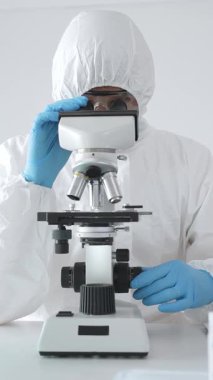 Tehlikeli madde giysisi giyen tıp virolojisti laboratuvarda mikroskopla çalışıyor. Yeni virüsler için potansiyel tedavileri araştırırken mavi sıvı örneklerini inceliyor.