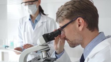 Bilimsel uzmanlar mikroskopla numuneleri inceliyor. Araştırma sırasında her ikisi de beyaz laboratuvar önlüğü giymiş koruyucu maske takan laboratuvar asistanı. Tıp ve Bilim.