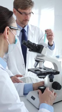 Koruyucu gözlük ve maske takan kadın bilim adamı, laboratuvarda tıbbi araştırma sırasında mikroskoptan bakıyor ve erkek meslektaşı arka planda çalışıyor. Tıp ve Bilim