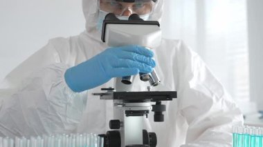 Tehlikeli madde giysisi giyen tıp virolojisti laboratuvarda mikroskopla çalışıyor. Yeni virüsler için potansiyel tedavileri araştırırken mavi sıvı örneklerini inceliyor.