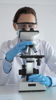 Koruyucu gözlük takan ve maske takan kadın bilim adamı mikroskop kullanarak laboratuvarda numuneleri analiz ediyor mavi sıvı test tüpleriyle çevrili yeni ilaç veya aşıları araştırıyor.
