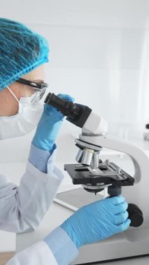 Mavi koruyucu giysiler giyen kadın bilim adamı mikroskop kullanarak laboratuvar ortamında örnekler inceleyerek bilimsel araştırma ve analiz yapıyor. Tıp ve bilim konsepti.