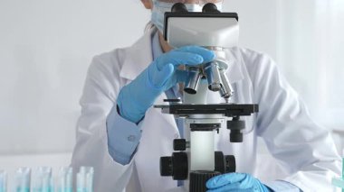 Mikroskobu ayarlayan, laboratuardaki örnekleri inceleyen, yeni ilaç veya kimyasal formülasyon araştırmaları yapan laboratuvar önlüğü giyen ve güvenlik için yüz maskesi takan bilim insanlarının eldiveni kapalı.
