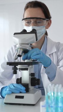 Koruyucu gözlük takan ve maske takan kadın bilim adamı mikroskop kullanarak laboratuvarda numuneleri analiz ediyor mavi sıvı test tüpleriyle çevrili yeni ilaç veya aşıları araştırıyor.