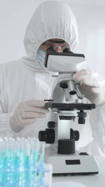 Laboratuvar ortamında araştırma yaparken mikroskop kullanarak koruyucu kıyafet giyen bir tıp bilimcisi. Test tüplerini mavi sıvıyla kapatın..