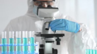 Laboratuvar ortamında araştırma yaparken mikroskop kullanarak koruyucu kıyafet giyen bir tıp bilimcisi. Test tüplerini mavi sıvıyla kapatın..