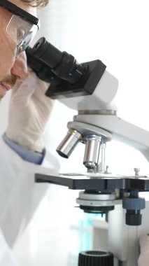 Erkek araştırmacının mikroskopla incelediği örnek, steril laboratuvar ortamında güvenlik gözlüğü takıyor, bilimsel cam eşyalar ile çevrili. Tıp, sağlık ve bilim konsepti.