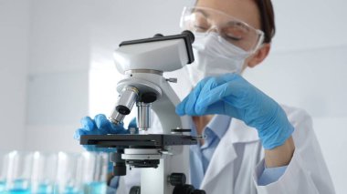 Koruyucu giysi giyen kadın bilim araştırmacısı mikroskobu kontrol ediyor, modern laboratuvar araştırma ortamındaki mavi test tüplerini analiz ediyor. Tıp ve bilim konsepti.