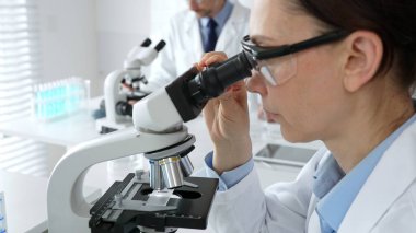 Koruyucu gözlük takan kadın bilim adamı mikroskop kullanarak modern laboratuvarda deney tüpleri ve arka planda erkek bilim adamı ile araştırma yapıyor. Tıp, sağlık ve bilim konsepti