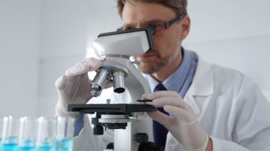 Laboratuvar önlüğü, gözlük ve beyaz koruyucu eldiven takan bilim araştırmacısı laboratuvar mikroskobunu dikkatlice kalibre ederek hassas tıbbi araştırma yapıyor. Tıp, sağlık ve bilim