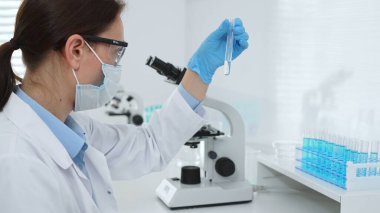 Koruyucu maske takan dişi araştırmacı ve mavi eldiven test tüpünde sıvıyı inceliyor, laboratuvarda mikroskop ve laboratuvar ekipmanlarının yanında oturuyor. Tıp, sağlık ve bilim konsepti.