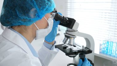 Maske takan ve mavi güvenlik donanımı takan kadın bir bilim adamı araştırma için mikroskop kullanırken erkek bir meslektaşı arka planda çalışıyor. Tıp, sağlık ve bilim konsepti.