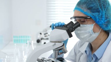 Koruyucu maske takan, gözlük takan ve mikroskop kullanarak laboratuvarda bilimsel araştırma yapan kadın bilim adamı. Tıp, sağlık ve bilim konsepti