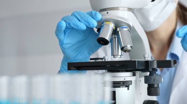 Mikroskobu ayarlayan eldivenli bilim adamlarına yakın çekim yapmak, laboratuvarda araştırma yapmak ve arka planda mavi sıvı içeren test tüpleri kullanmak. Tıp, sağlık ve bilim konsepti.