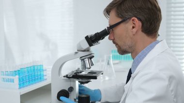 Laboratuvar araştırmacısı mavi sıvı örneklerini analiz ediyor, mikroskop altında detaylı bilimsel araştırma yaparken koruyucu giysi giyiyor. Tıp, sağlık ve bilim konsepti