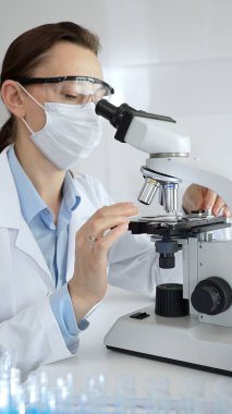 Maskeli profesyonel kadın bilim adamı mikroskop ile numune inceleyerek steril laboratuvar ortamında meslektaşlarıyla birlikte araştırma yapıyor. Tıp, sağlık ve bilim konsepti.