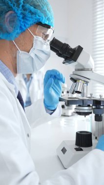 Koruyucu maske, şapka ve eldiven takan kıdemli erkek bilim adamı modern bir laboratuvarda mikroskop altında örnekleri inceliyor. Tıp, sağlık ve bilim konsepti.