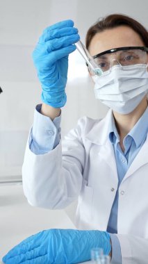 Steril laboratuvar ortamında tıbbi deney sırasında koruyucu giysi giyerken mavi sıvıyı analiz eden deney tüpü tutan kadın araştırmacı. Tıp ve bilim konsepti.
