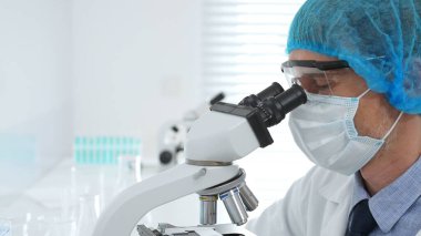 Kıdemli bilim adamı mikroskopla numune inceliyor, steril laboratuvar ortamında tıbbi araştırma yaparken koruyucu giysiler giyiyor. Tıp, sağlık ve bilim konsepti