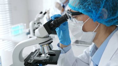 Maske takan ve mavi güvenlik donanımı takan kadın bir bilim adamı araştırma için mikroskop kullanırken erkek bir meslektaşı arka planda çalışıyor. Tıp, sağlık ve bilim konsepti.