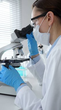 Maske takan ve mavi eldiven giyen kadın bilim adamı mikroskop kullanarak arka planda çalışan bir meslektaşla modern laboratuvarda bilimsel araştırmalar yapıyor. Tıp, sağlık ve bilim konsepti.