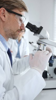 Koruyucu gözlük takan bilim adamı mikroskop altında araştırma ve analiz yapıyor. Arka planda test tüpleri ile modern bir laboratuvarda. Tıp, sağlık ve bilim konsepti