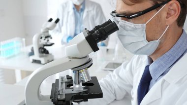 Mikroskop kullanarak maske takan ve eldiven takan bilim adamlarına yakın çekim, modern laboratuvarda araştırmalar yürütmek ve arka planda çalışmak. Tıp, sağlık ve bilim konsepti