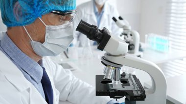 Koruyucu maske, şapka ve eldiven takan kıdemli erkek bilim adamı modern bir laboratuvarda mikroskop altında örnekleri inceliyor. Tıp, sağlık ve bilim konsepti.