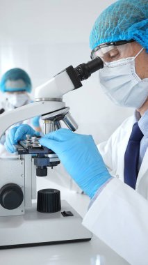 Koruyucu maske, şapka ve eldiven takan kıdemli bir bilim adamı modern bir laboratuvarda mikroskop altında örnekleri inceliyor. Tıp, sağlık ve bilim konsepti.