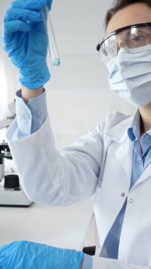 Steril laboratuvar ortamında tıbbi deney sırasında koruyucu giysi giyerken mavi sıvıyı analiz eden deney tüpü tutan kadın araştırmacı. Tıp ve bilim konsepti.