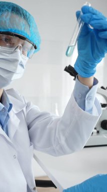 Steril laboratuvar ortamında tıbbi araştırma sırasında mikroskop yakınında mavi sıvı numunesini inceleyen koruyucu ekipman giyen bilim adamları. Tıp, sağlık ve bilim konsepti