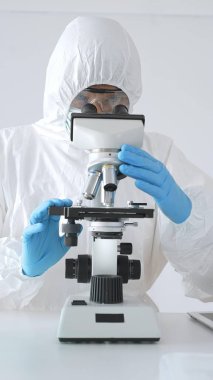 Laboratuvar ortamında araştırma yaparken mikroskop kullanarak koruyucu kıyafet giyen bir tıp bilimcisi. Test tüplerini mavi sıvıyla kapatın..