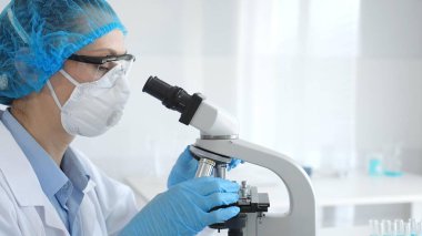 Kadın araştırmacı mikroskop ile numune inceliyor, steril laboratuvar içinde çalışma yerini çevreleyen cam şişelerle tıbbi deney yapıyor. Tıp ve Bilim.