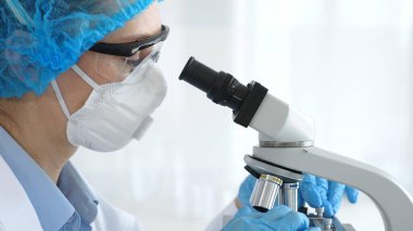 Mavi renkli koruyucu teçhizatlı kadın araştırmacı mikroskop ile numune inceliyor, laboratuvar içinde cam şişelerle laboratuvar içinde deney yapıyor. Tıp ve Bilim.