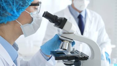 Mikroskop altında numuneleri incelerken mavi koruyucu giysili kadın bilim adamlarının yakın plan çekimlerinde laboratuvar asistanları arka planda cam parçaları incelerler. Tıp konsepti.