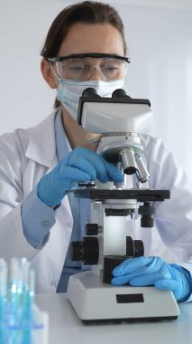 Mikroskop kaydırağını inceleyen kadın bilim adamı, laboratuarda koruyucu giysiler giyiyor, mavi renkli test tüpleriyle farmasötik buluşları araştırıyor..