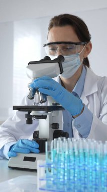 Mikroskop kaydırağını inceleyen kadın bilim adamı, laboratuarda koruyucu giysiler giyiyor, mavi renkli test tüpleriyle farmasötik buluşları araştırıyor..