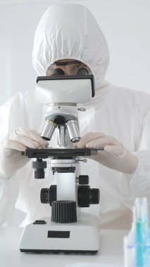 Tehlikeli madde giysisi giyen tıp virolojisti laboratuvarda mikroskopla çalışıyor. Yeni virüsler için potansiyel tedavileri araştırırken mavi sıvı örneklerini inceliyor.