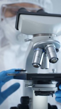 Tehlikeli madde giysisi, maskesi ve eldiveni olan bir tıp virologu bir laboratuvar araştırma virüsünün mikroskobunda özenle çalışıyor. Sağlık ve Bilim Konsepti.