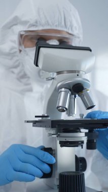 Tehlikeli madde giysisi, maskesi ve mavi eldivenli bir erkek virüs uzmanı laboratuvar araştırmalarında mikroskop başında özenle çalışıyor. Tıp, bilim konsepti.